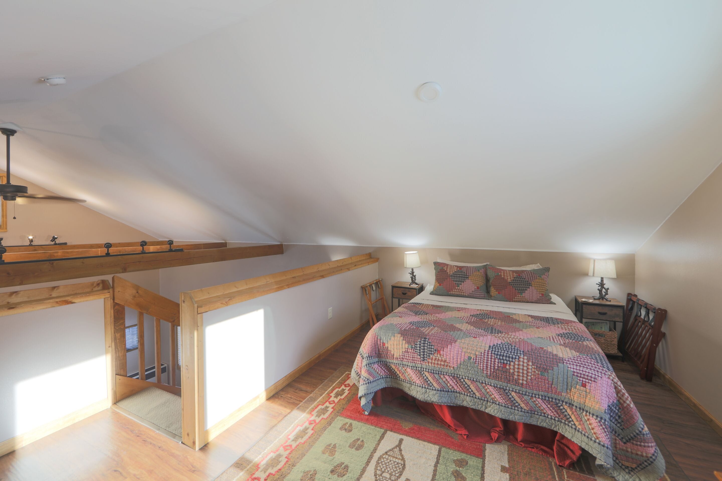 A bedroom with a wooden ceiling, a bed with a patterned bedspread, a nightstand with a lamp, and a wooden chair.