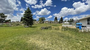 A grassy field with a large evergreen tree, a white picket fence, and a blue barrel near a building. - Callie's Cottage- Cottage stay between Bryce and Zion (Hatch)