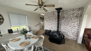 A dining area with a white table, white chairs, a ceiling fan, a brick-patterned wall, a black wood stove, and a staircase leading to another room. - Callie's Cottage- Cottage stay between Bryce and Zion (Hatch)