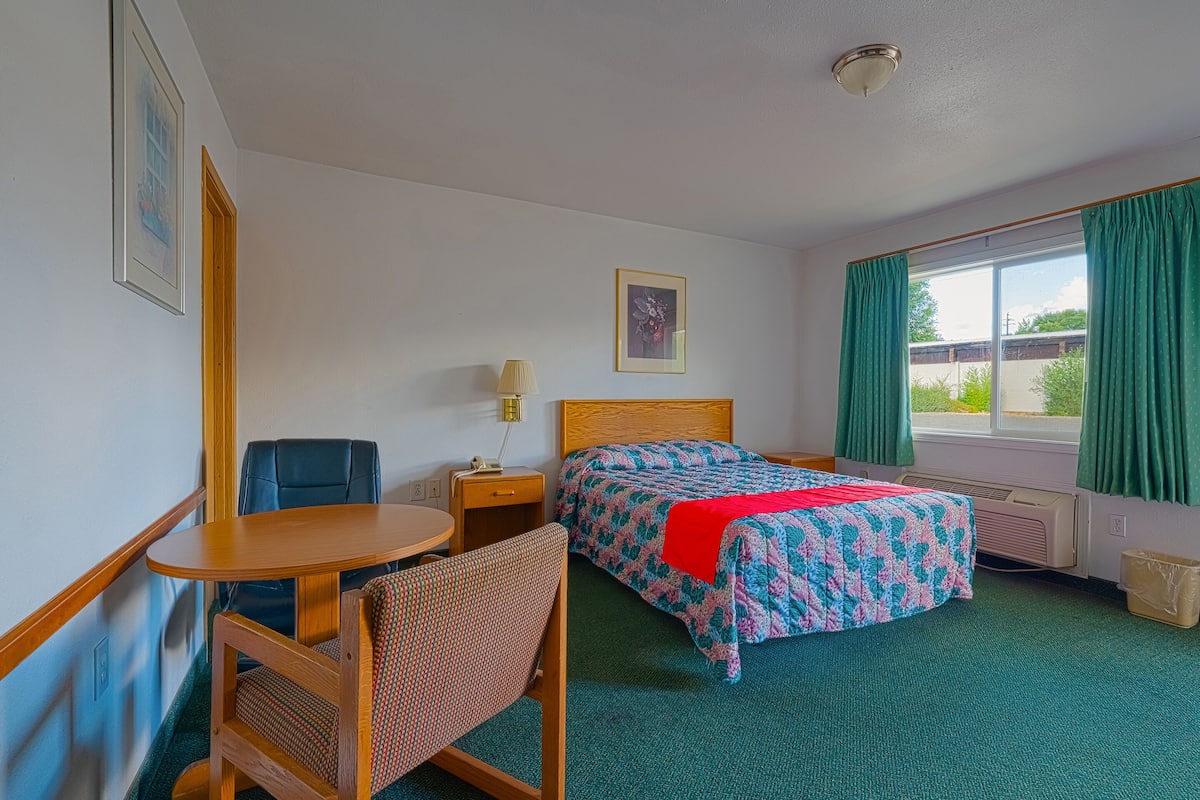 Ein Hotelzimmer mit Bett, Schreibtisch, Stuhl, einem Fenster mit Vorhängen und einem Bild an der Wand.