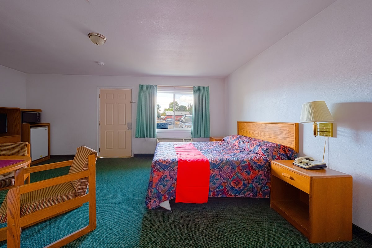 Ein Hotelzimmer mit einem Bett, einem Schreibtisch mit Lampe, einem Sessel, einem kleinen Tisch und einer Tür zu einem Balkon.