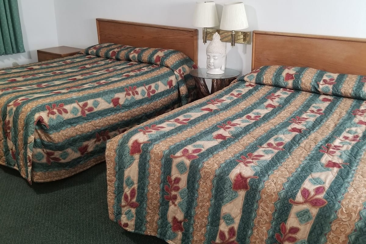 Ein Hotelzimmer mit zwei Betten, beide mit gemusterten Bettdecken. Zwischen den Betten befindet sich ein Nachttisch mit zwei Wandlampen.