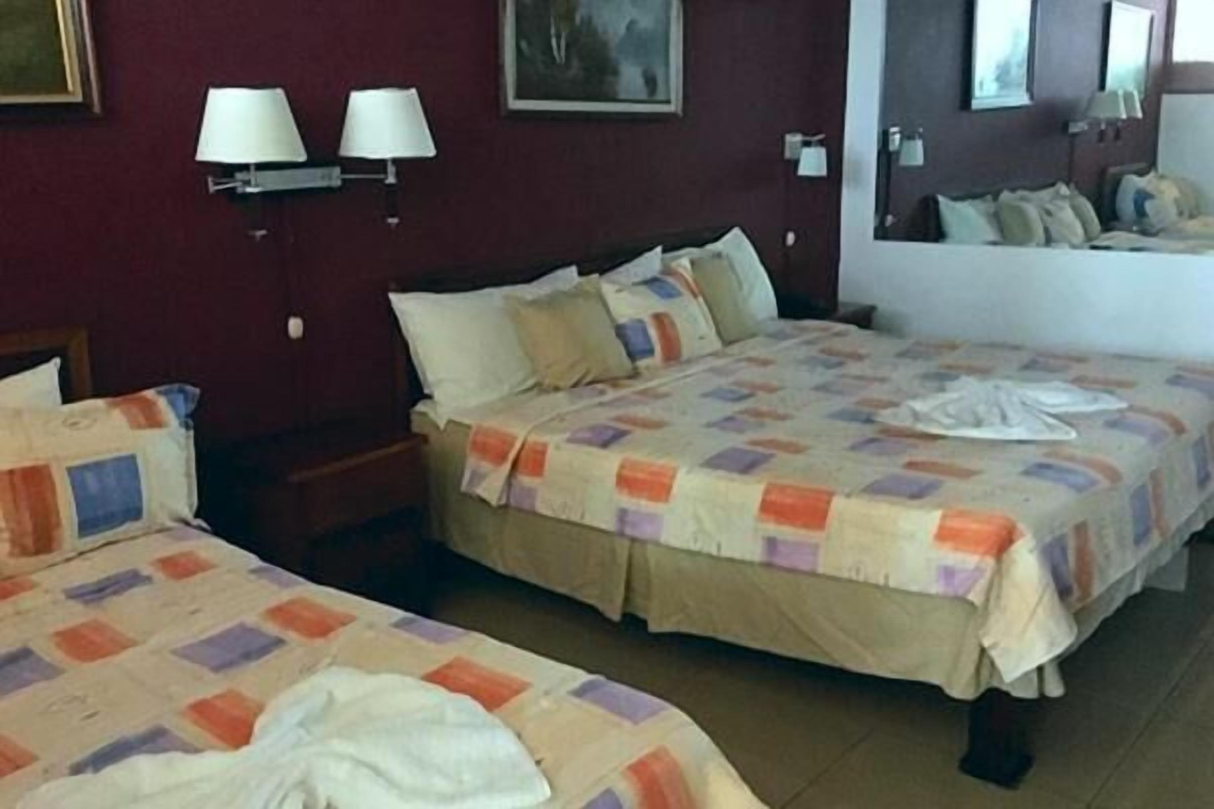 Una habitación de hotel con dos camas, cada una con una colcha acolchada y almohadas. Hay lámparas fijadas a la pared y un cuadro enmarcado.