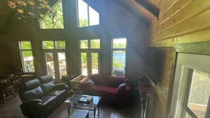 A spacious living room with a leather sofa, a glass coffee table, and a fireplace. Large windows provide views of the outdoors.