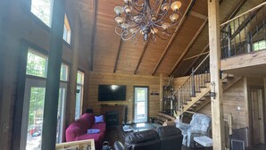 A spacious living room with a large chandelier, wooden walls, a red sofa, a black leather couch, and a staircase leading to an upper level.