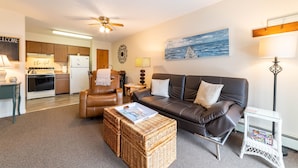 A living room with a leather sofa, a wicker ottoman, a glass coffee table, and a small side table with magazines. There is a kitchen in the background with wooden cabinets and a refrigerator. - Awesome in Walloon Village (Walloon Lake)