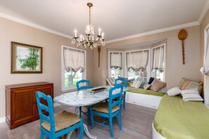Un coin repas avec une table ronde, des chaises bleues et un lustre. À gauche, on trouve un banc près de la fenêtre agrémenté de coussins et un meuble en bois.