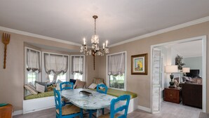 A dining area with a round table, blue chairs, a chandelier, and a window seat with cushions.