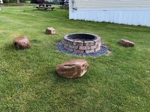 A fire pit surrounded by rocks in a grassy yard with a picnic table and a building in the background.