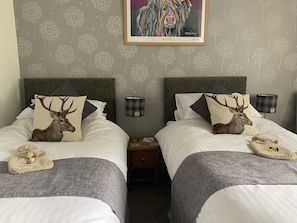 A hotel room with two beds, each with a pillow featuring a deer. There is a framed artwork above the beds, a bedside lamp, and a small nightstand between the beds. - Morlea Bed & Breakfast (Inverness)
