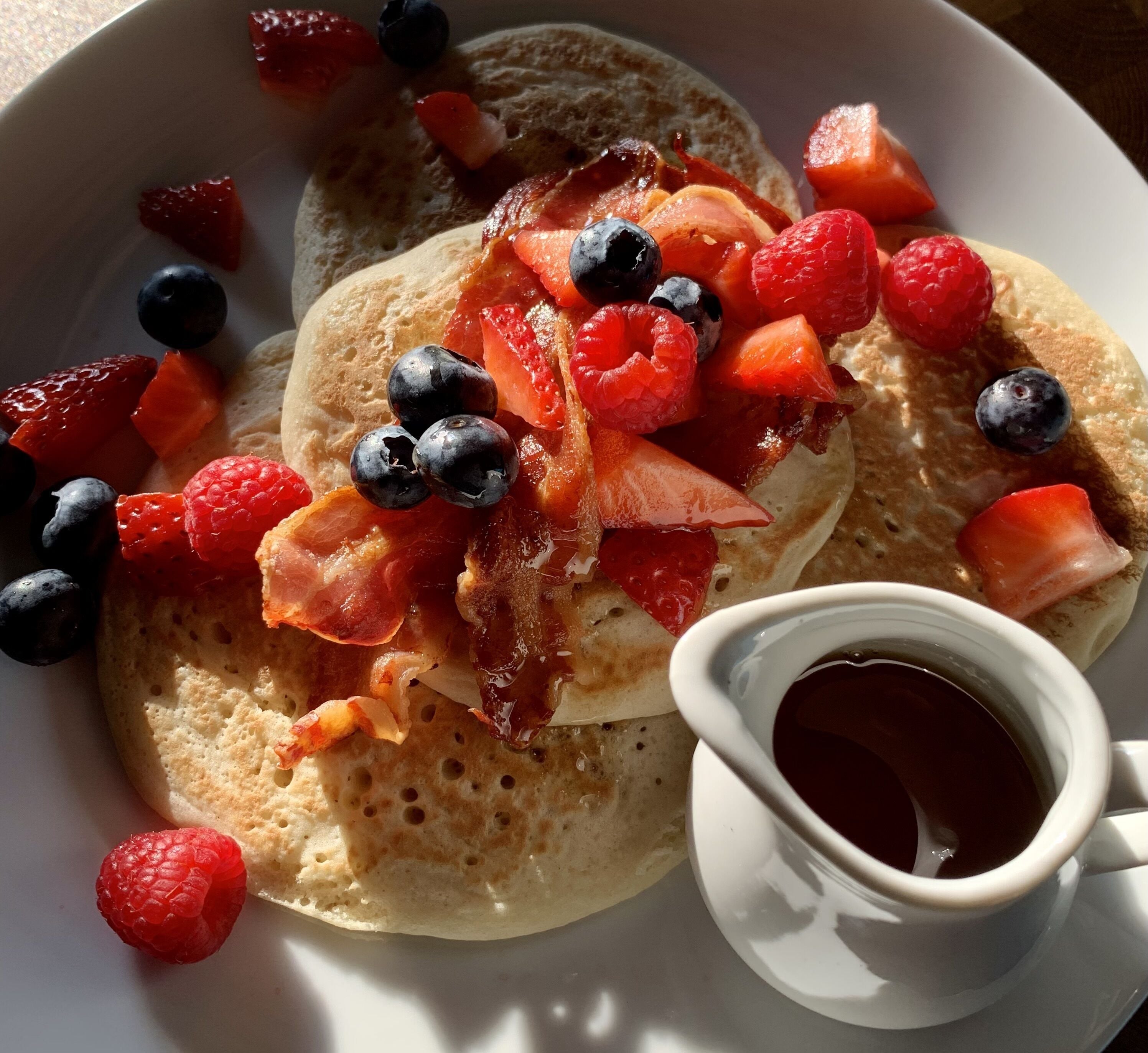Un piatto di pancake guarnito con pancetta, mirtilli, lamponi e fragole, accompagnato da una tazza di sciroppo.