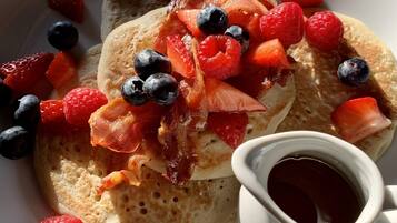 Un piatto di pancake guarnito con pancetta, mirtilli, lamponi e fragole, accompagnato da una tazza di sciroppo.