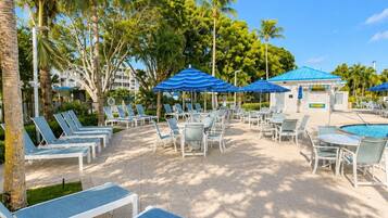 A resort pool area with sun loungers, umbrellas, and palm trees.