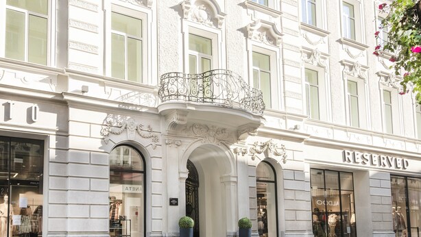 Un bâtiment doté d’éléments architecturaux décoratifs, notamment un balcon orné d’une balustrade en fer forgé, ainsi que des devantures de magasins avec des enseignes.