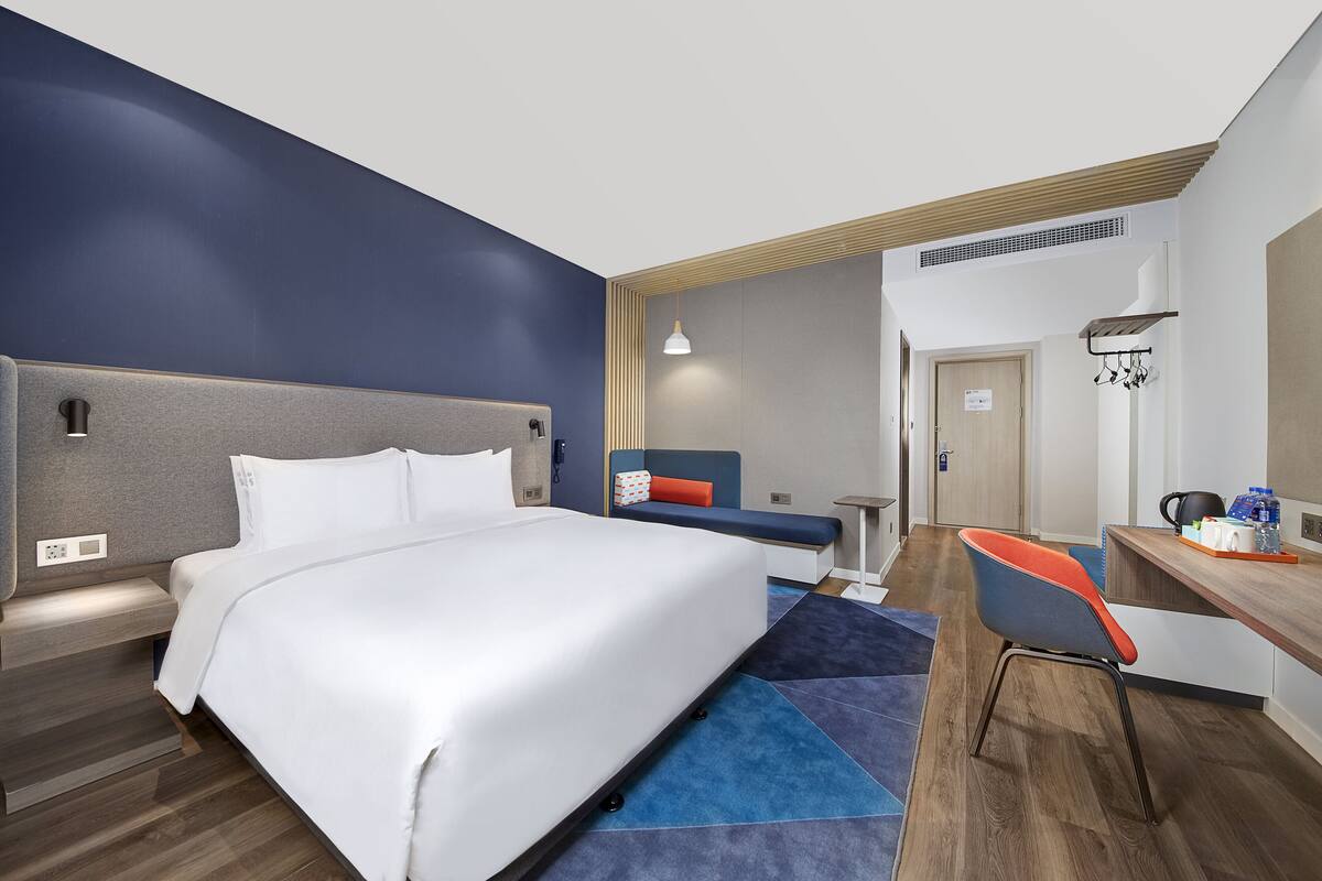 A modern hotel room with a large bed, a desk with a chair, a small seating area, and a kitchenette.