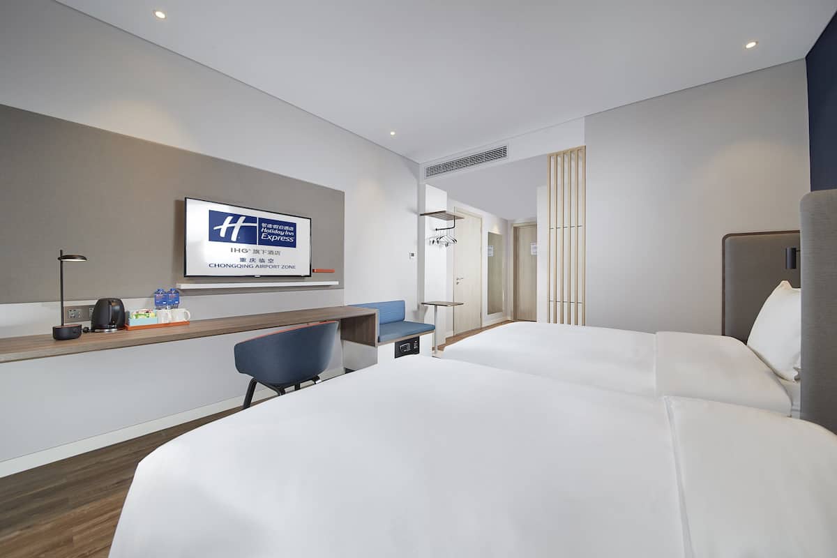 A hotel room with two beds, a desk with a chair, a TV mounted on the wall, and a small seating area.