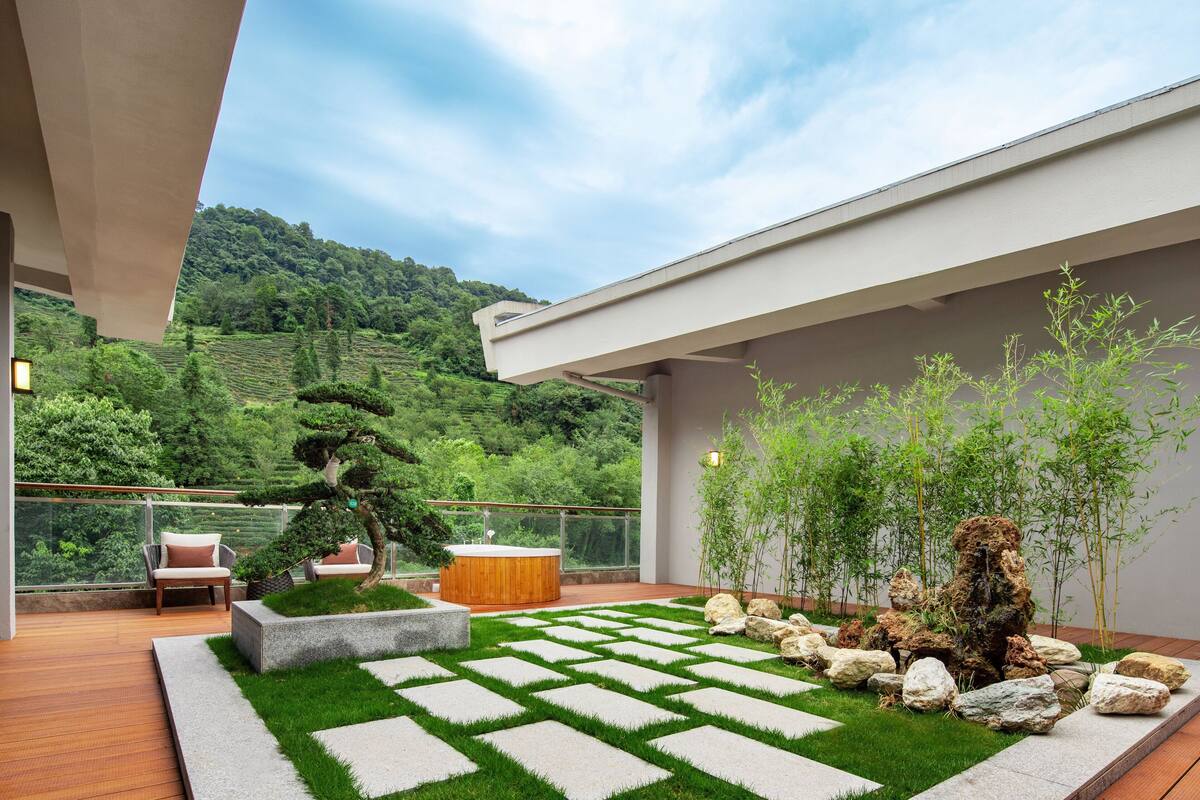 A modern outdoor patio with a wooden deck, a seating area with a cushioned chair, a wooden hot tub, and a garden with a stone pathway and greenery.
