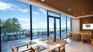Un espace de restauration moderne dans un hôtel, avec de grandes fenêtres, des meubles en bois et une vue sur l’extérieur.