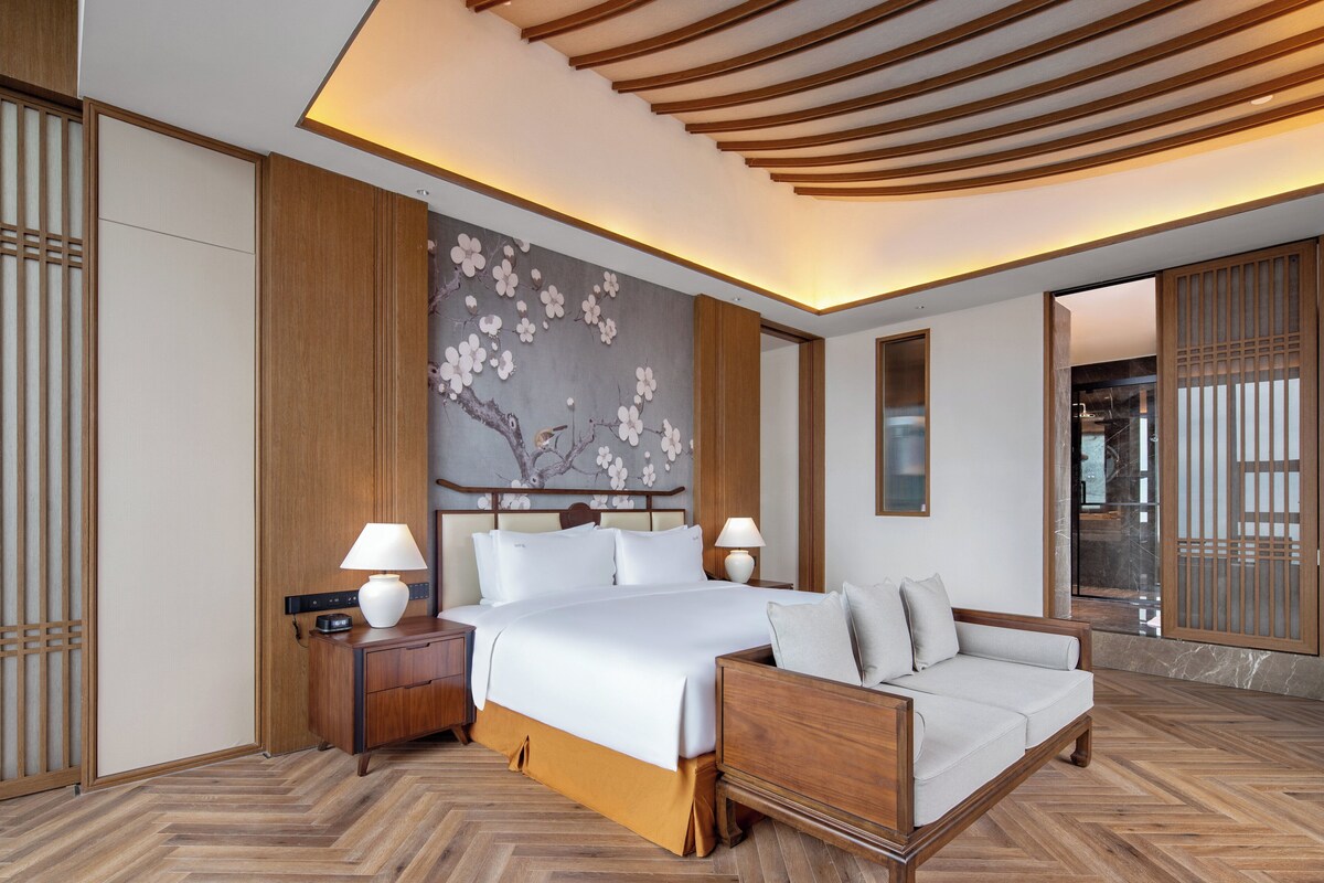 A modern hotel room with a large bed, a sofa, wooden flooring, and a decorative wall with a tree and flowers.
