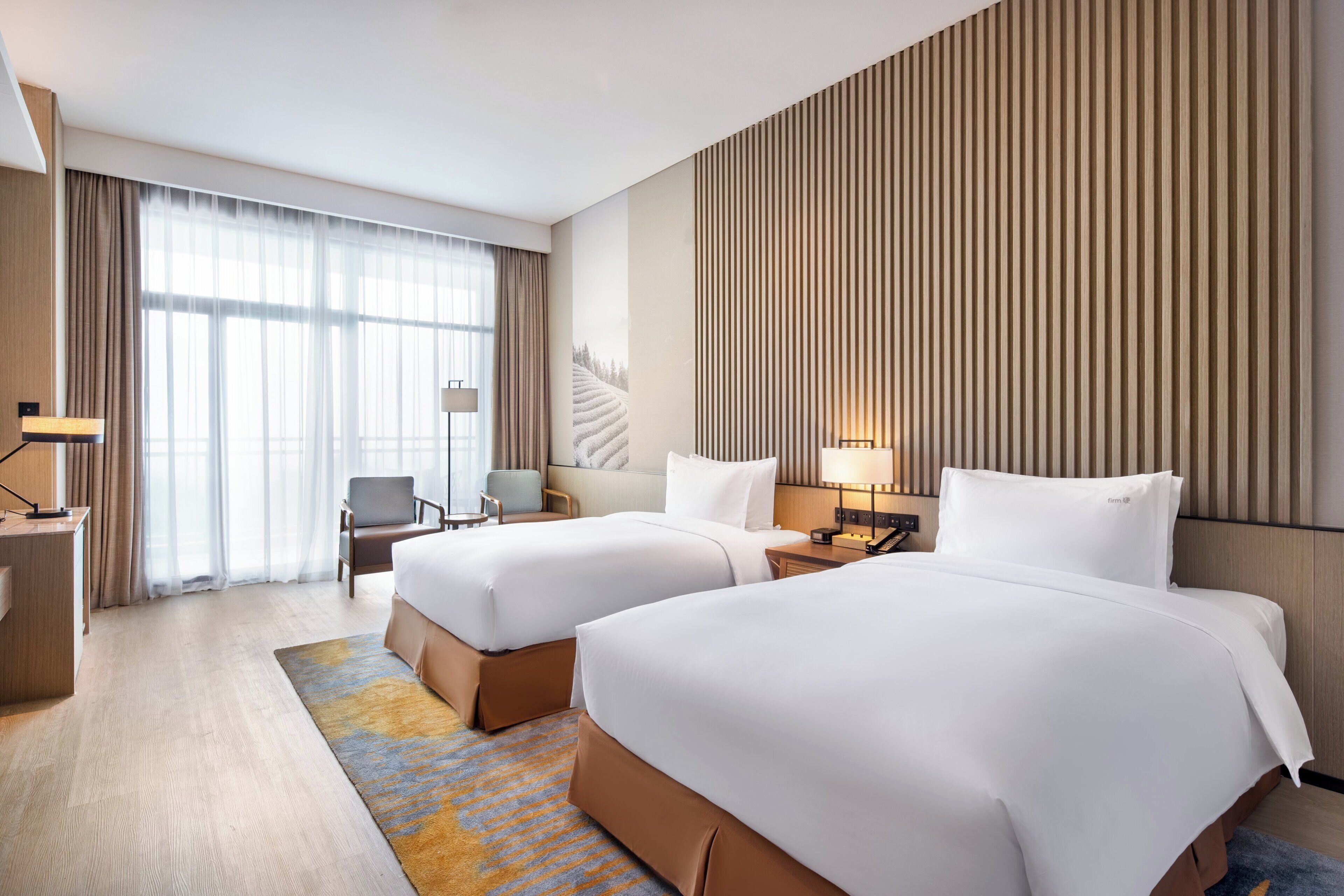 A hotel room with two beds, a desk, a chair, a lamp, and a large window with curtains.