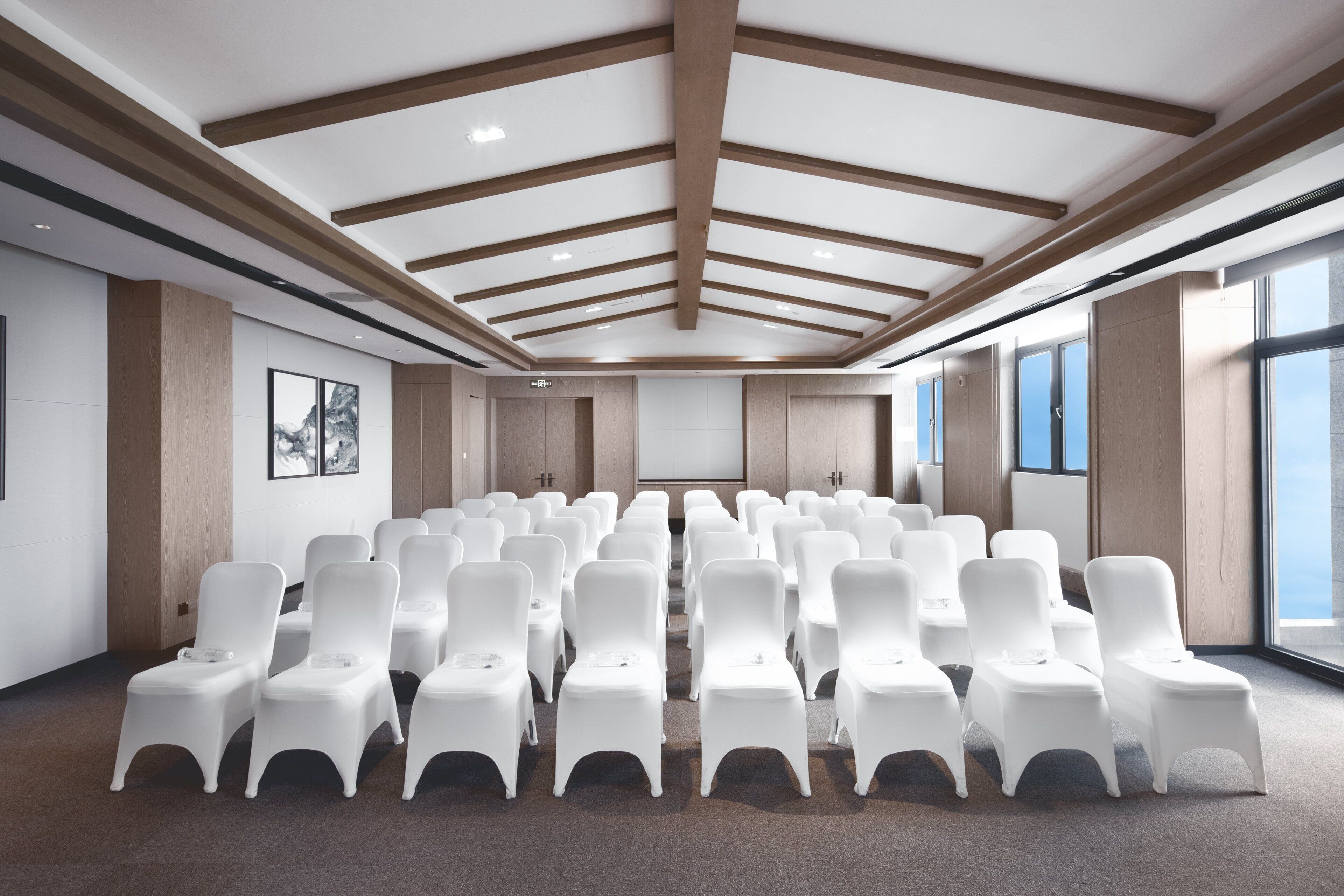 Un salón de conferencias con filas de sillas blancas, vigas de madera en el techo y grandes ventanales a un lado.