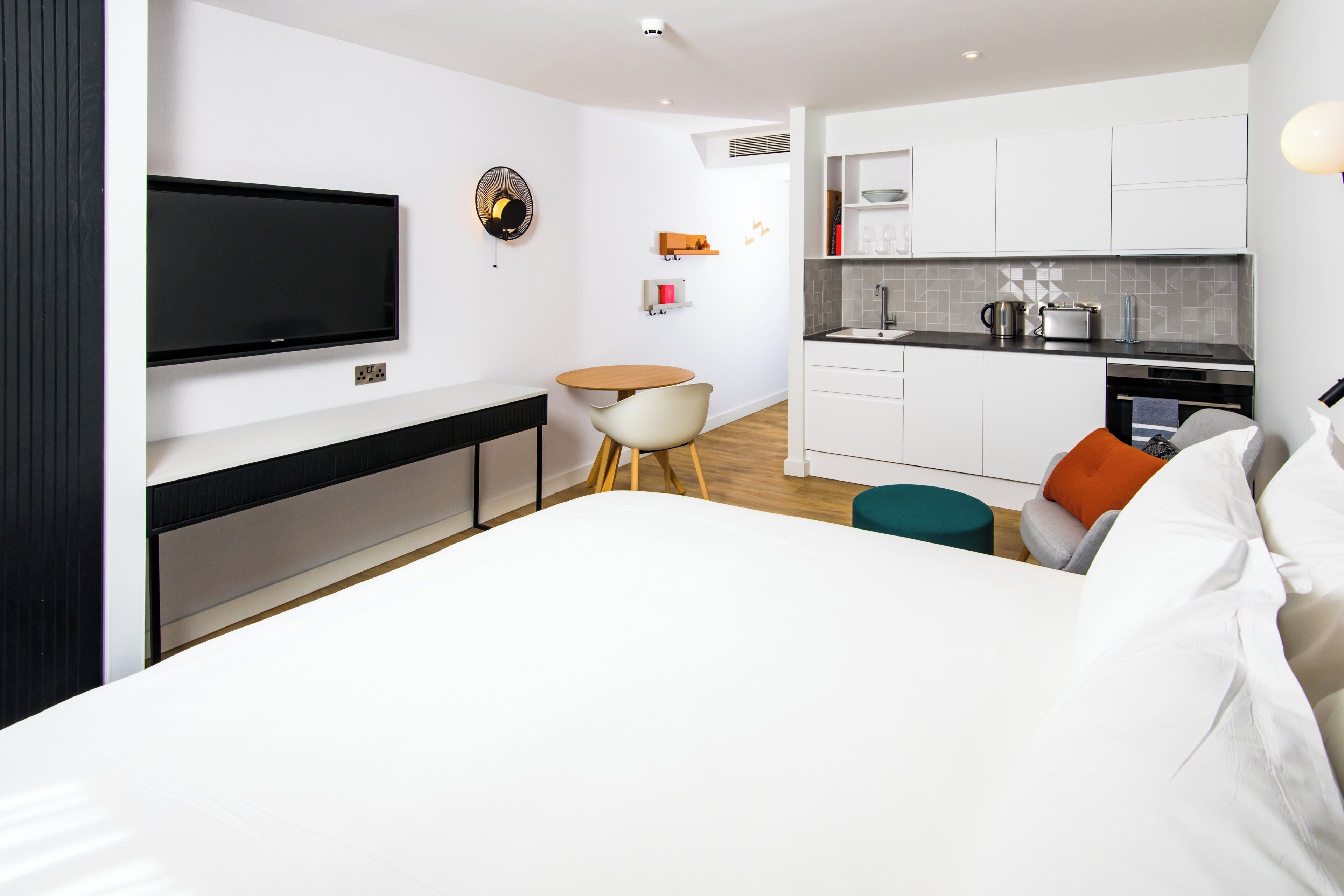 A modern hotel room with a large bed, a TV mounted on the wall, a small round table, and a kitchenette with a sink and a toaster.