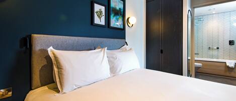 A modern hotel room with a large bed, a nightstand, a wall-mounted light, and a bathroom visible through an open door.
