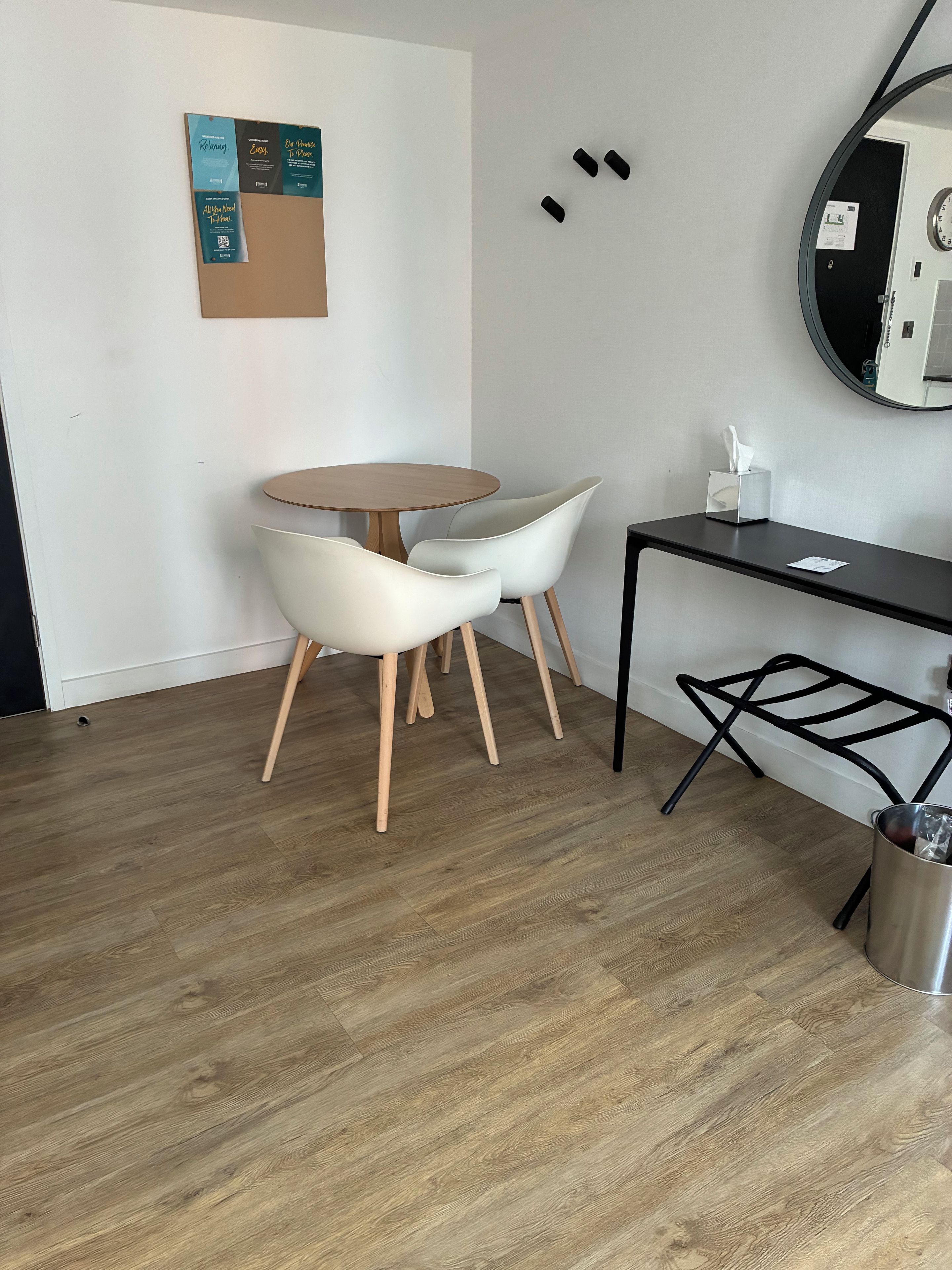 Quarto moderno com uma mesa redonda, duas cadeiras brancas, uma escrivaninha de metal preto e uma lixeira.