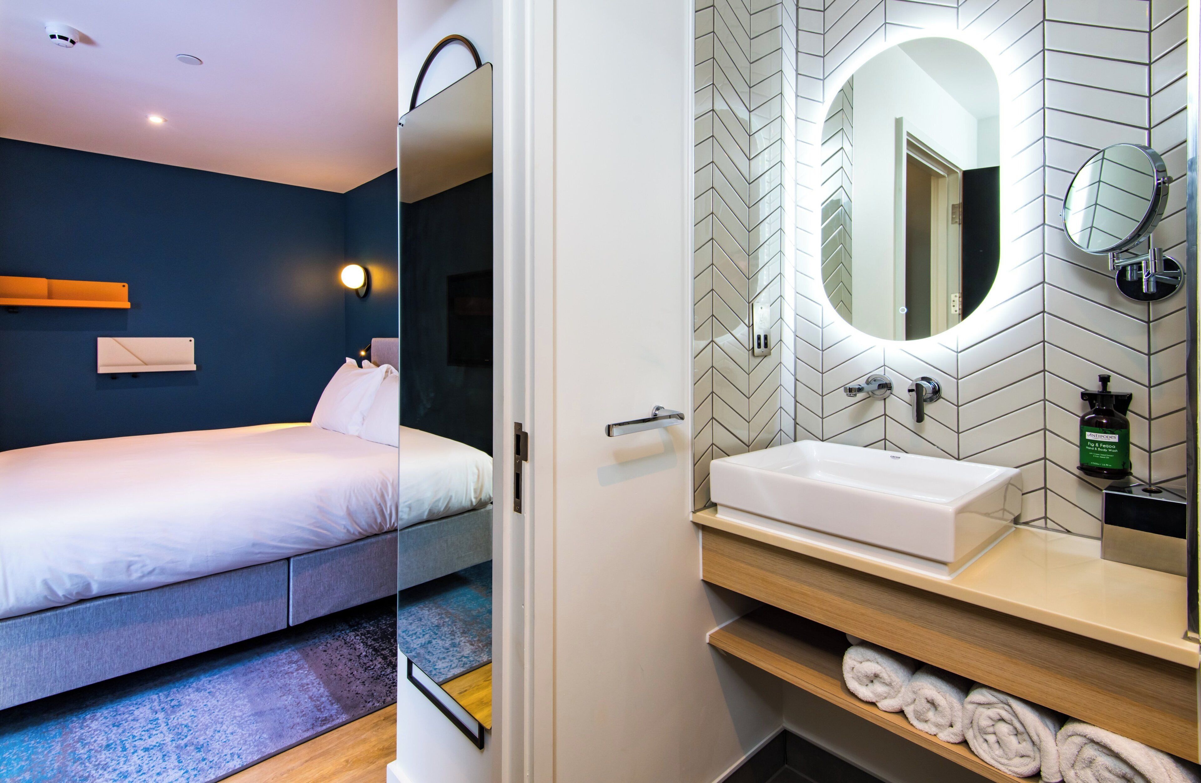 Une chambre d’hôtel moderne équipée d’un lit, d’une table de chevet et d’une salle de bain avec un lavabo, un miroir et un porte-serviettes.