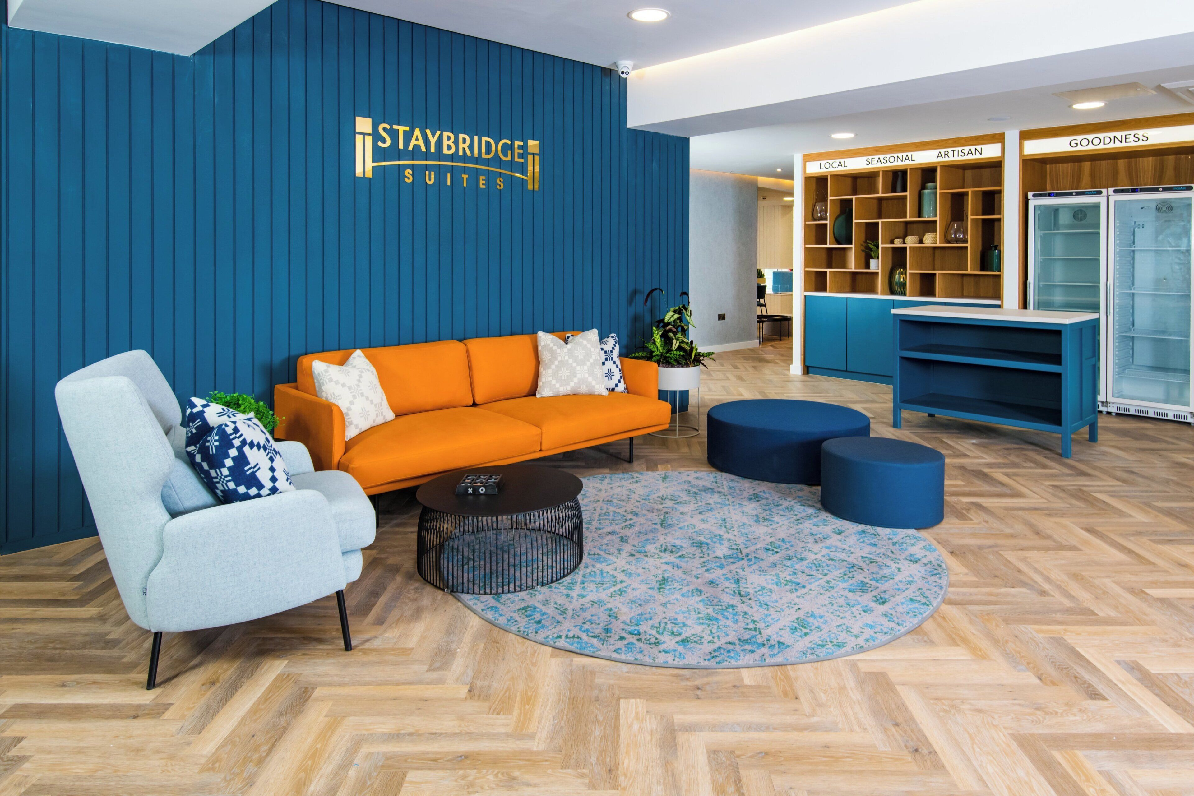 A modern hotel lobby with a blue wall, a Staybridge Suites sign, a sofa, a coffee table, and a bookshelf labeled 'GOODNESS'.