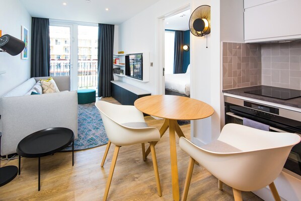 A modern living room with a sofa, two white chairs, a round wooden table, and a kitchen area with a stove and oven. - Staybridge Suites Cardiff by IHG (Cardiff)