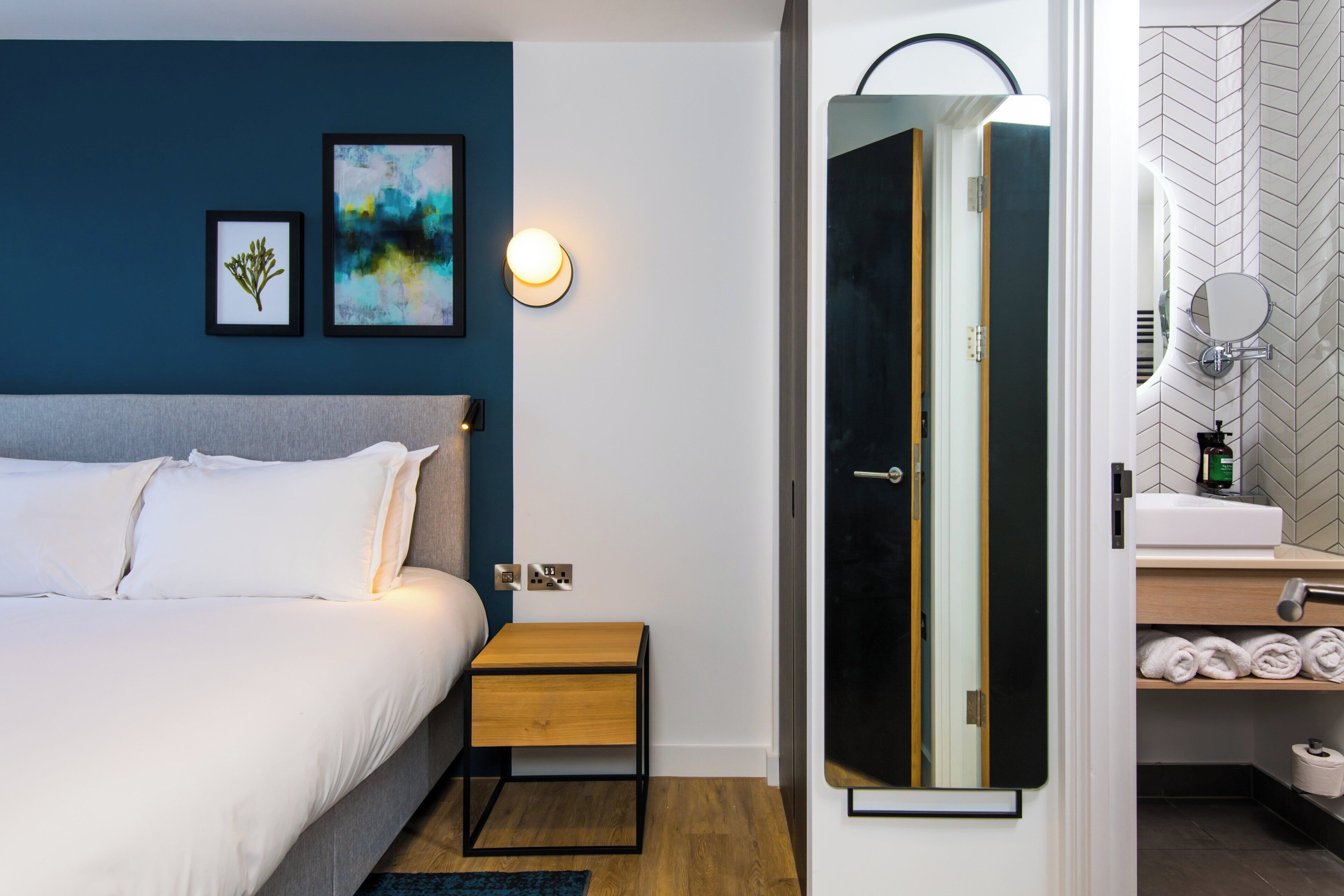 A bedroom with a bed, bedside table, wall art, and a bathroom with a mirror, sink, and towel rack.