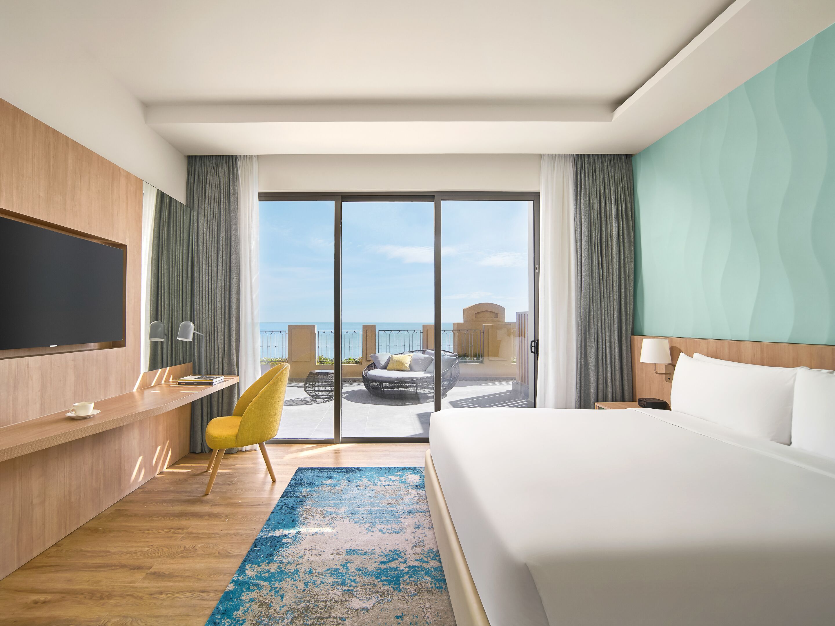 Habitación de hotel con una cama grande, un escritorio con silla, un televisor montado en la pared y un balcón con vista al mar.
