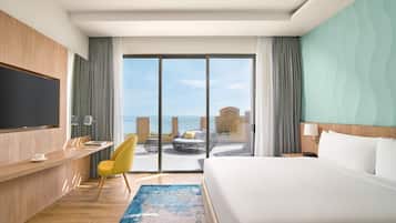 A hotel room with a large bed, a desk with a chair, a TV mounted on the wall, and a balcony with a view of the sea.