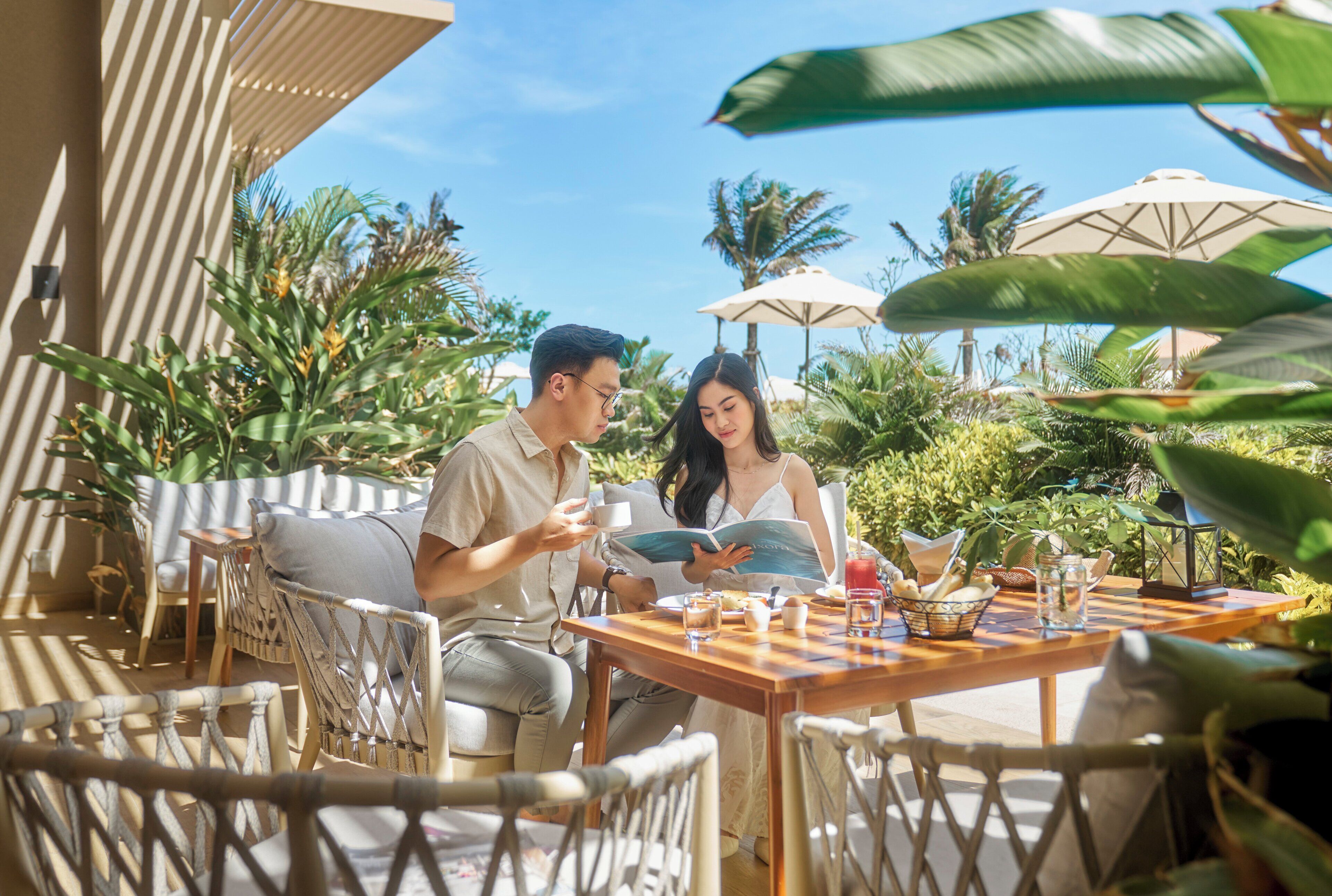 A couple sitting at an outdoor table with a menu, surrounded by wicker chairs and umbrellas, with lush greenery in the background.