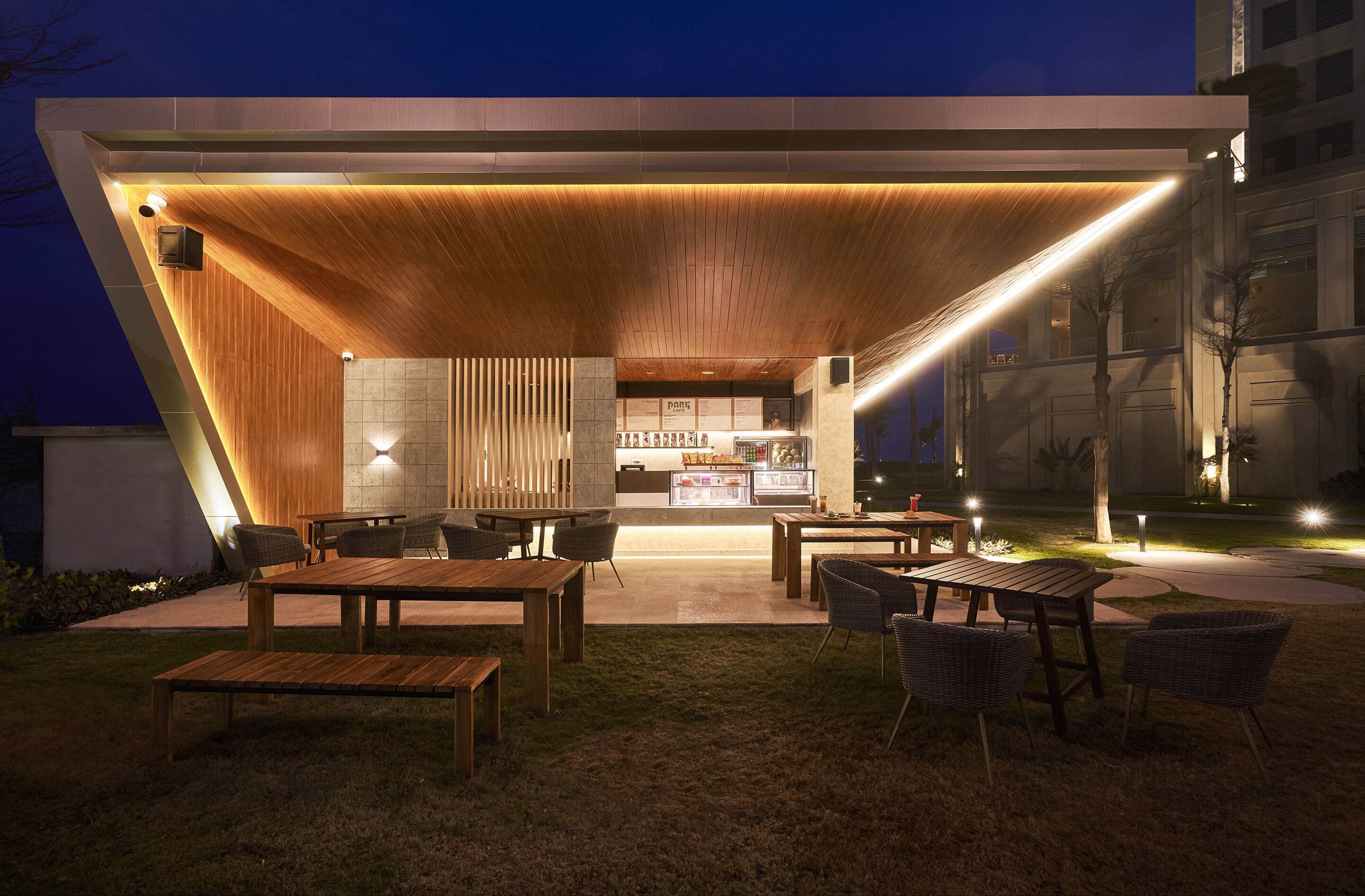 An outdoor dining area with wooden tables and chairs, a bar, and a well-lit entrance.