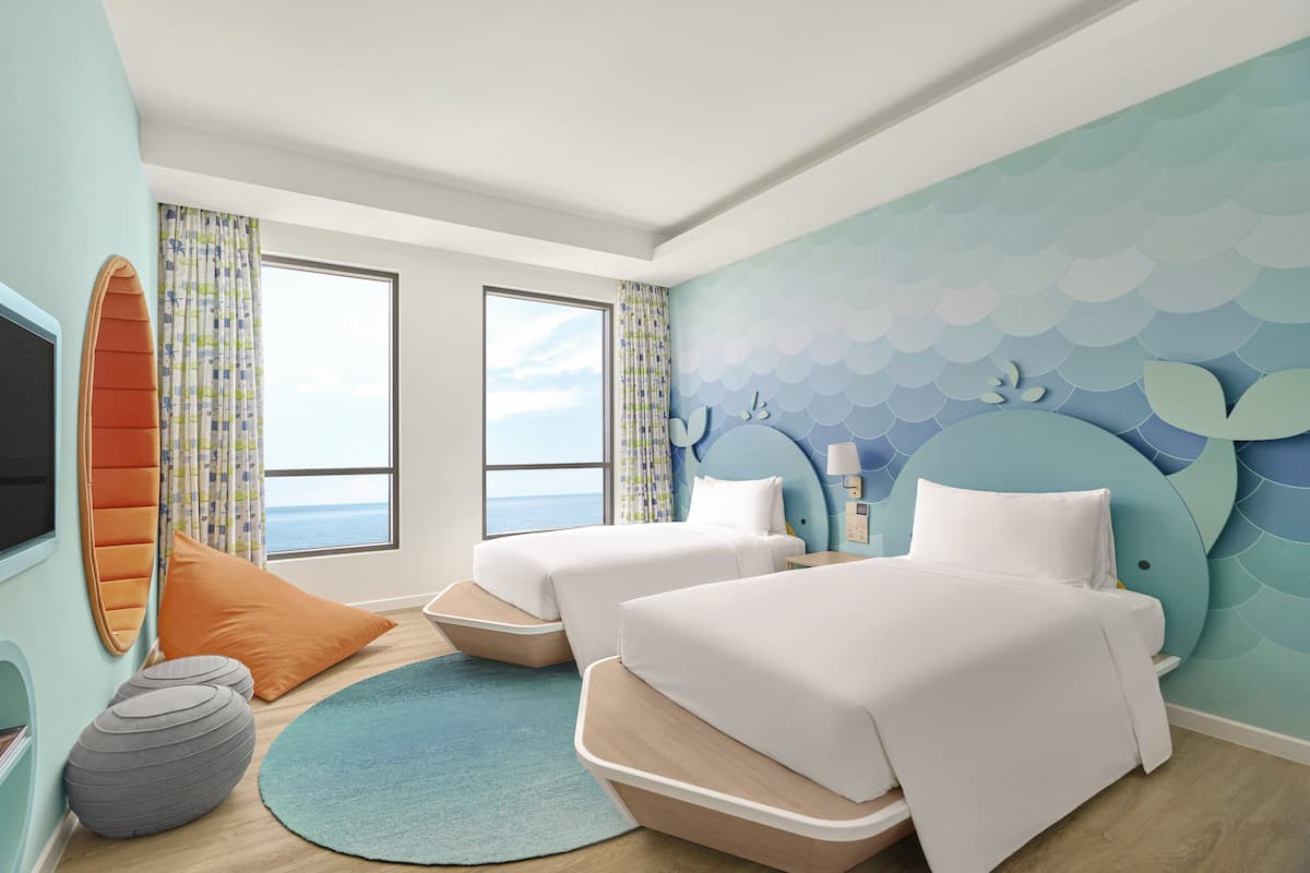 A hotel room with two beds, a large window, a surfboard decoration, and a sea-themed wall.