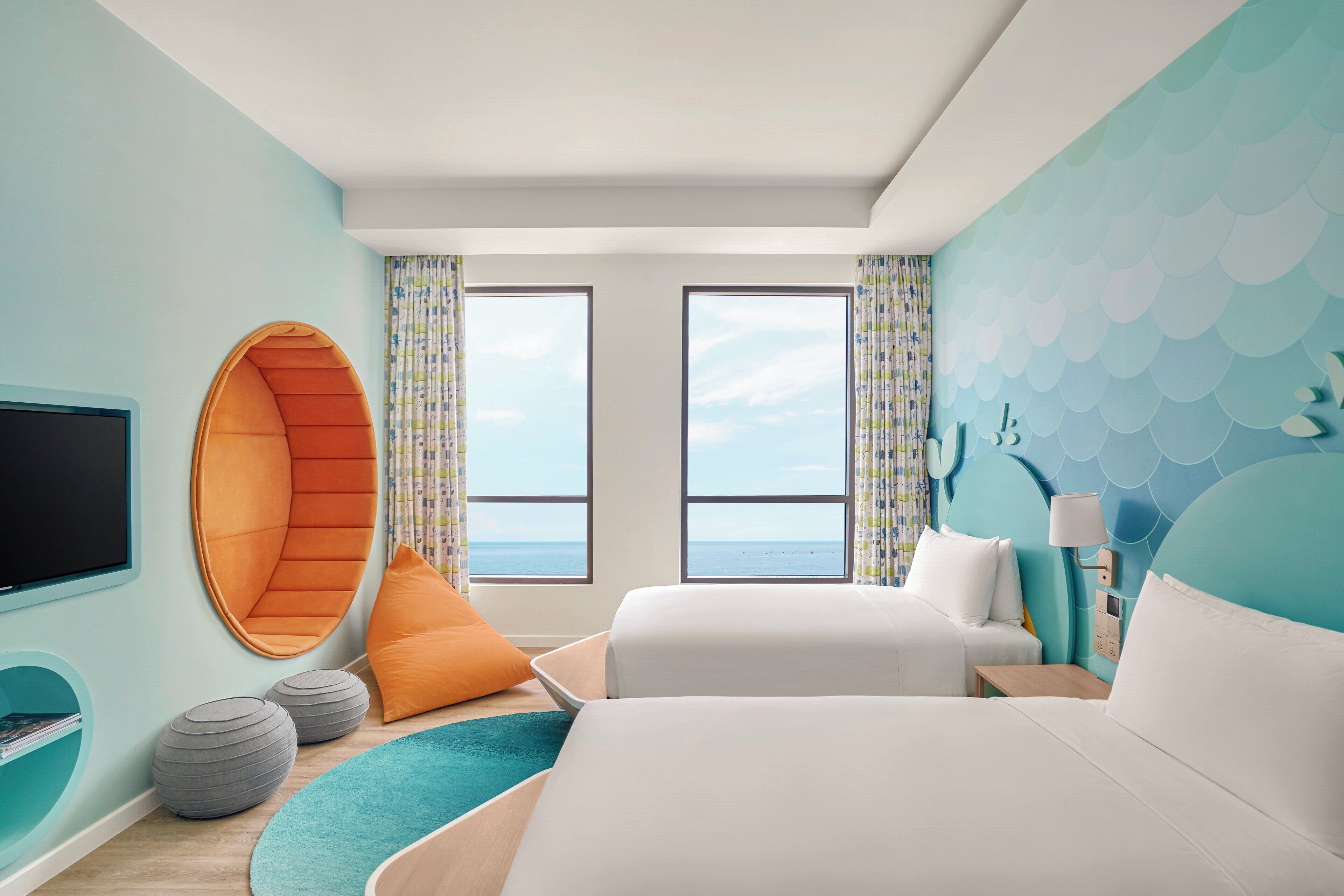 A modern hotel room with a large bed, a curved seating area, two windows with ocean views, and a wall-mounted TV.