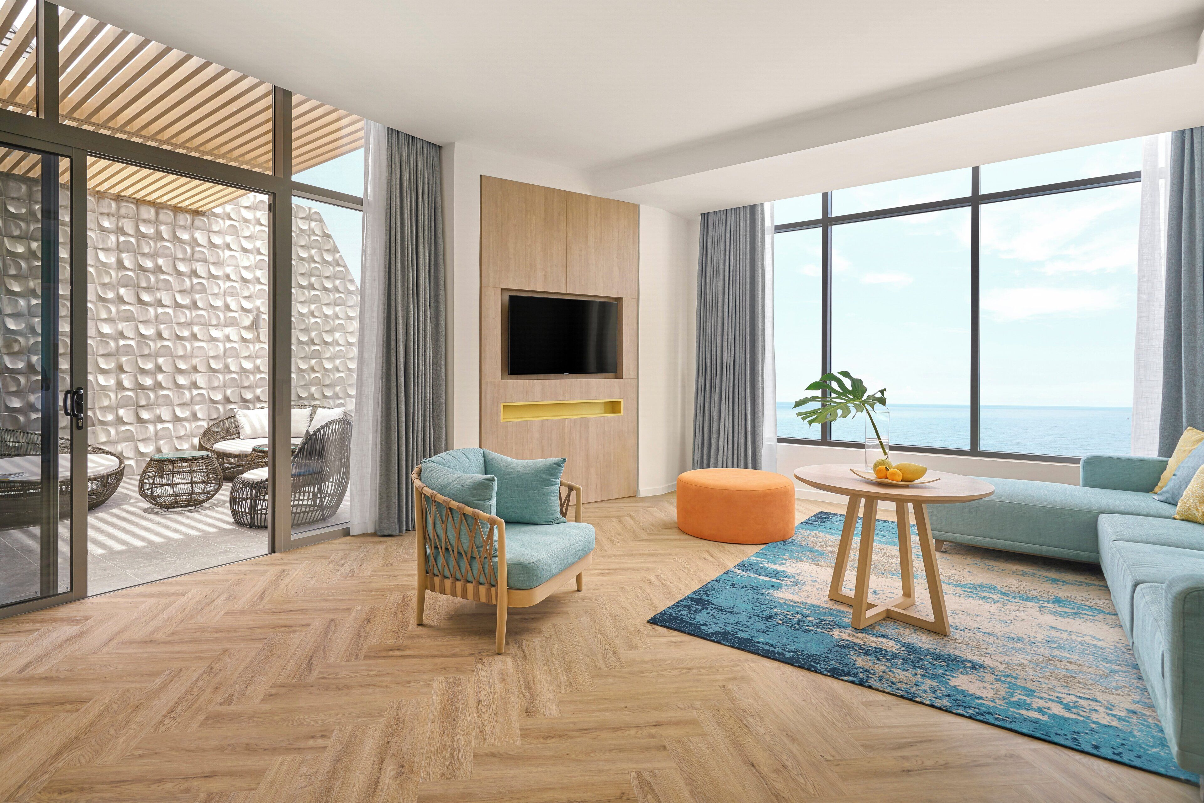 A modern living room with a large window, a flat-screen TV, wicker furniture, and a view of the sea.