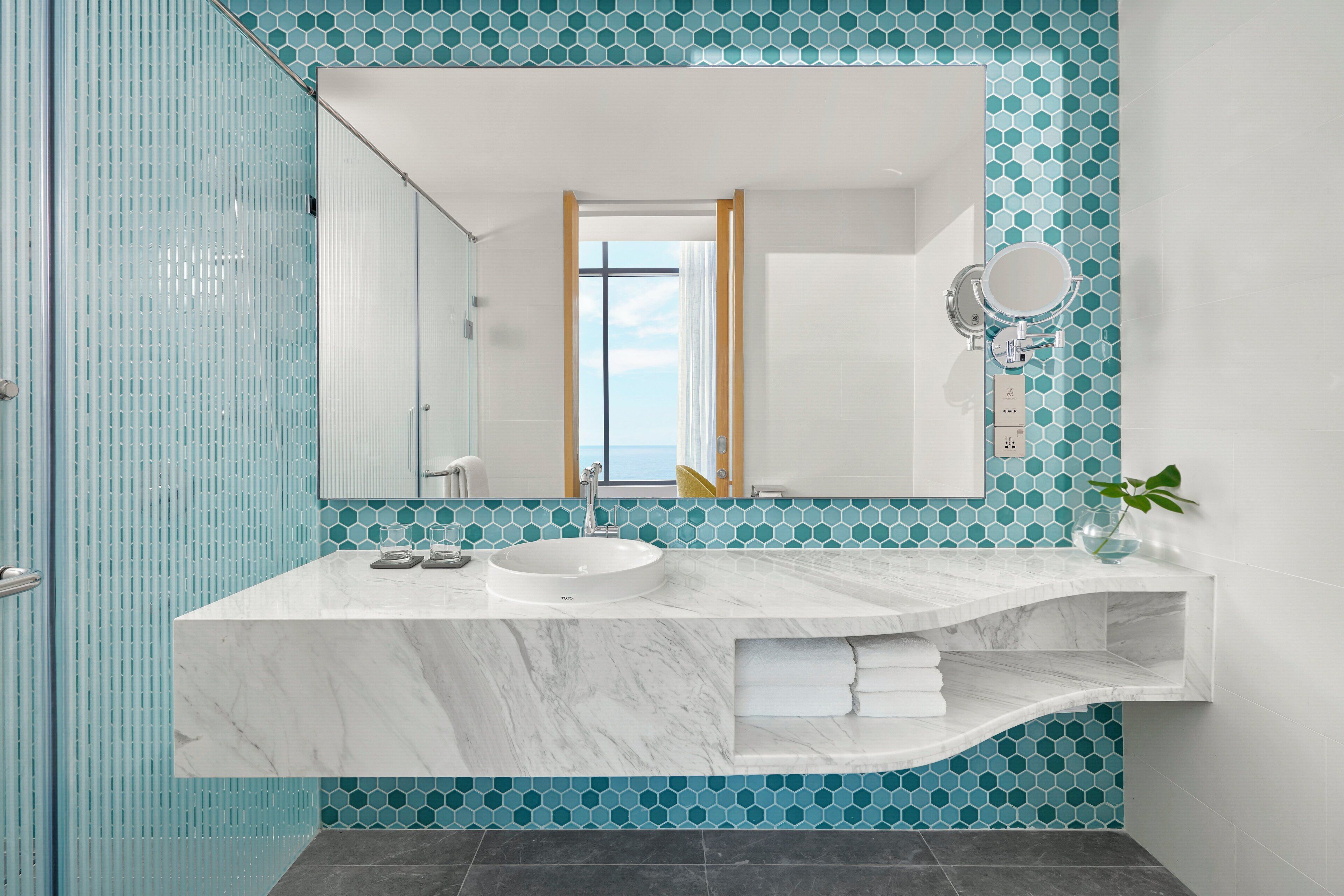 Une salle de bain avec un lavabo en marbre blanc, un grand miroir et une vue sur la mer par une fenêtre.
