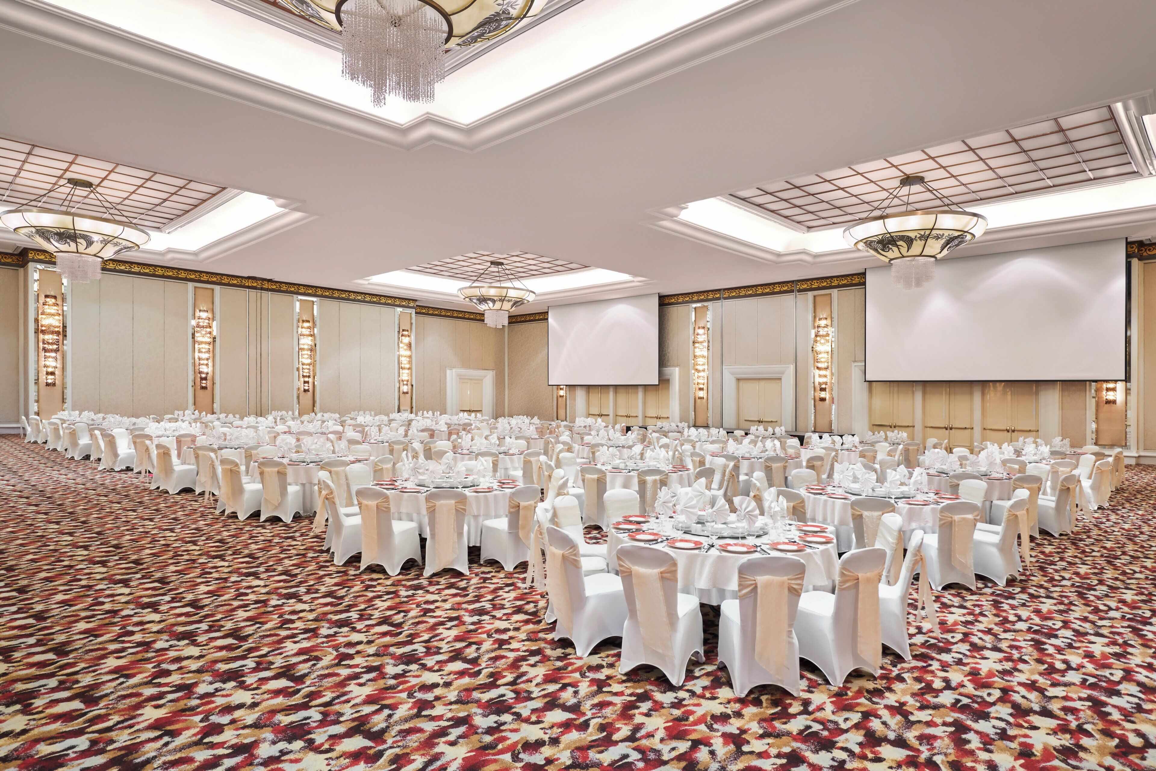 A large banquet hall with rows of round tables set for dining, white chairs, and chandeliers.