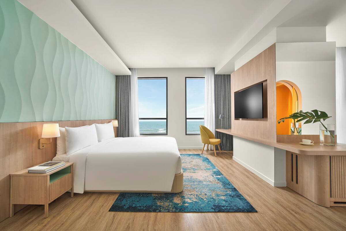 A modern hotel room with a large bed, a desk, a TV, and a view of the sea.