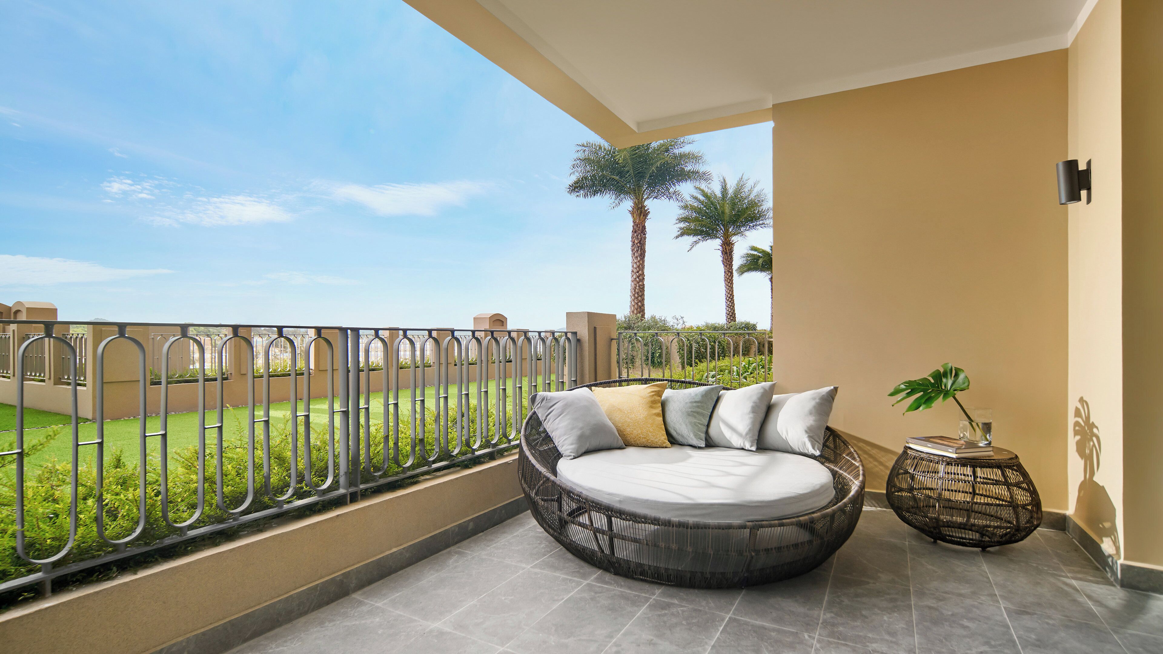 A balcony with a round wicker sofa, a small side table with a plant, and a view of a green landscape with palm trees.