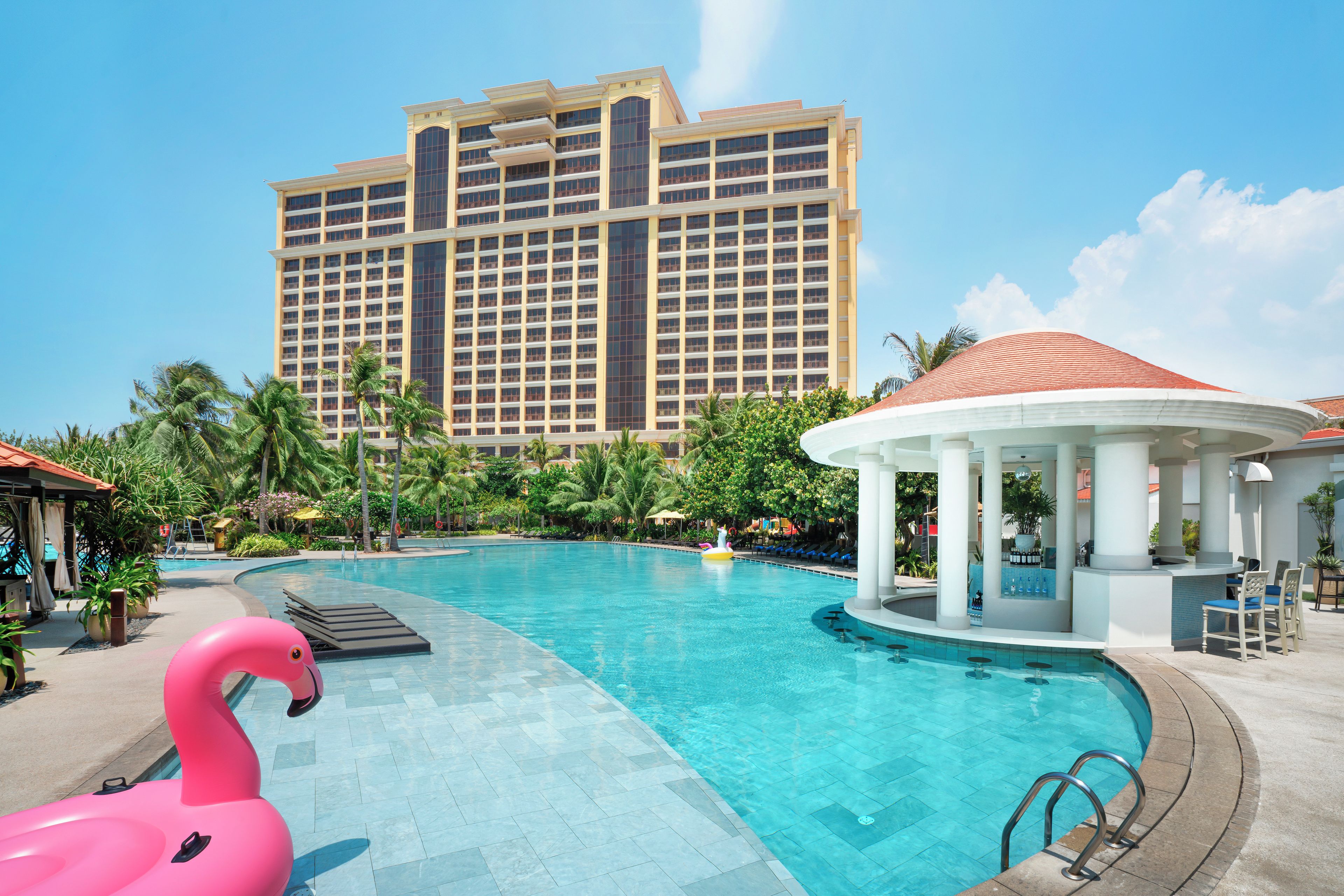 A swimming pool with a pink flamingo float, a gazebo, and a large hotel building in the background.