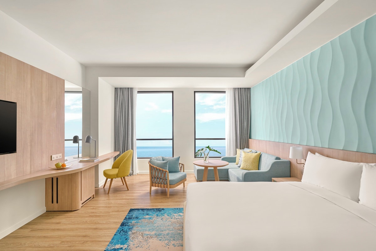 A modern hotel room with a large bed, a desk, a chair, and a sofa. There are two windows with curtains, and a view of the sea.