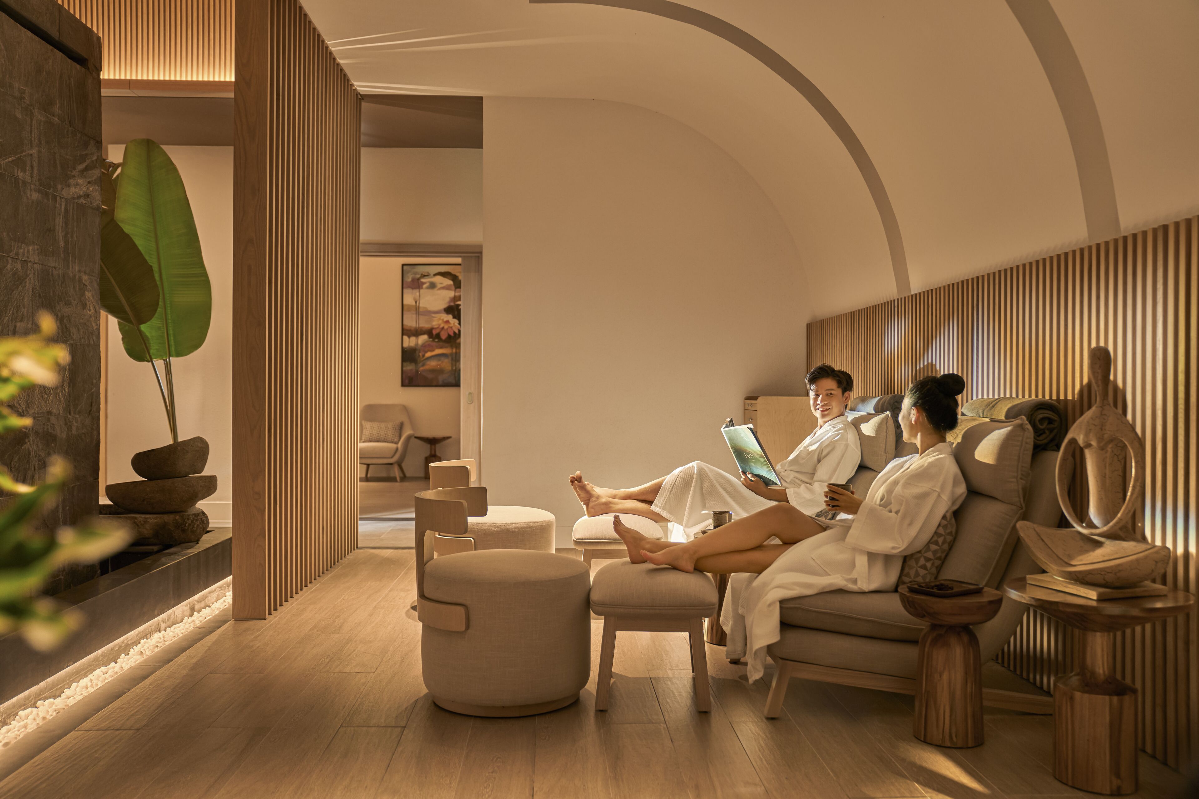 A modern spa room with wooden flooring, a large plant, and a couple relaxing on a sofa.