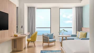 A hotel room with a bed, a desk, a chair, a sofa, and large windows with a view of the sea.