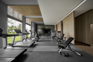 Un gimnasio moderno con cintas de correr, elípticas y bancos de pesas, dispuesto en un corredor amplio con ventanas grandes y vistas al verdor.