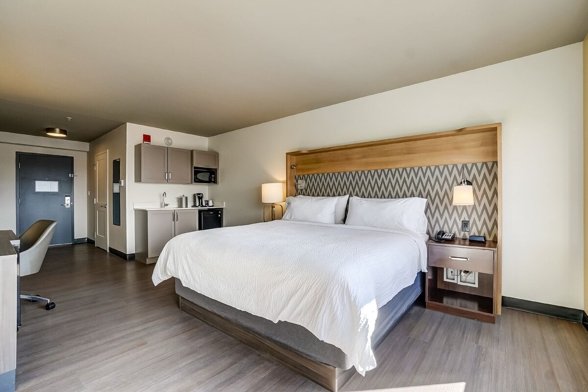 A hotel room with a large bed, bedside lamps, a wooden headboard, a desk with a chair, and a kitchenette.