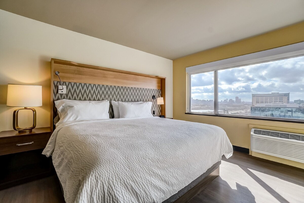 A hotel room with a large bed, two bedside lamps, a view of the city through a large window, and a modern headboard.